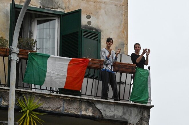 14 mars 2020. À Rome, de jeunes gens sur leur balcon applaudissent en chantant. Un appel lancé sur les réseaux sociaux a suggéré aux Italiens de chanter tous à la même heure pour sortir de l’isolement lié aux précautions face au Covid-19. © Andreas Solaro / AFP