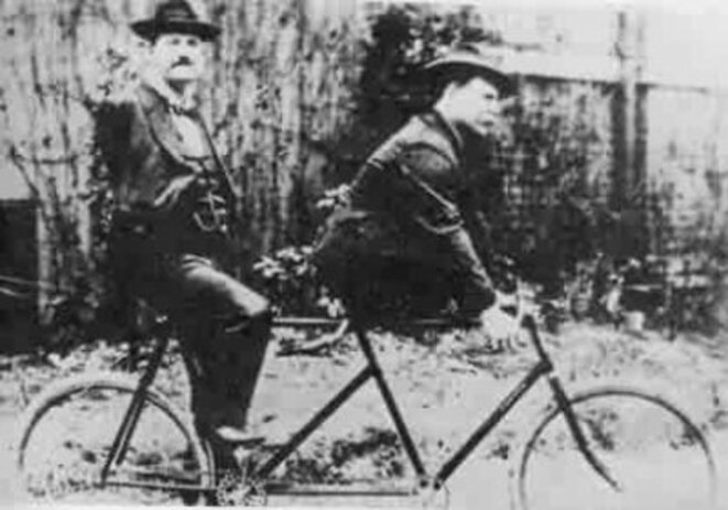 photo ancienne en noir et blanc avec un tandem, le cycliste arrière n'a pas de bras, le cycliste avant n'a pas de jambes